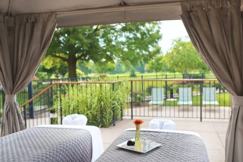 A serene outdoor spa setting with two massage tables, a flower vase, and a view of lush greenery and lounge chairs.