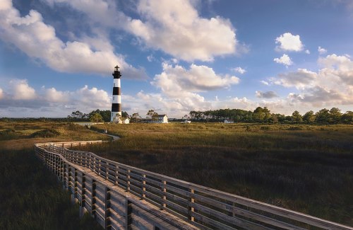 A lighthouse stands tall against a blue sky, surrounded by grassy marshland and a wooden walkway leading to it.
