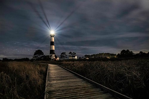 A lighthouse beams light at night, surrounded by tall grass and a wooden walkway leading to it under a starry sky.
