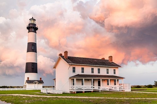 A black-and-white striped lighthouse beside a white house, set against a colorful sunset and dramatic clouds.