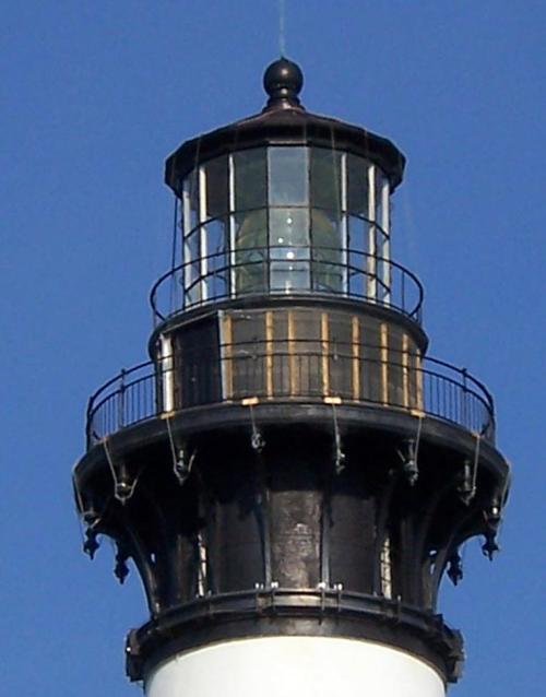 Close-up of a lighthouse lantern room with a glass dome and metal railing against a clear blue sky.