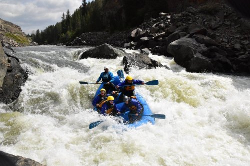 A group of six people white-water rafting on a turbulent river, surrounded by rocky banks and lush greenery.