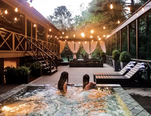 Two women relax in a hot tub on a deck adorned with string lights, surrounded by greenery and a cozy outdoor setting.