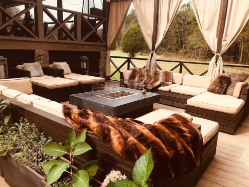 Cozy outdoor seating area with plush sofas, a fire pit, and decorative throws, surrounded by greenery.