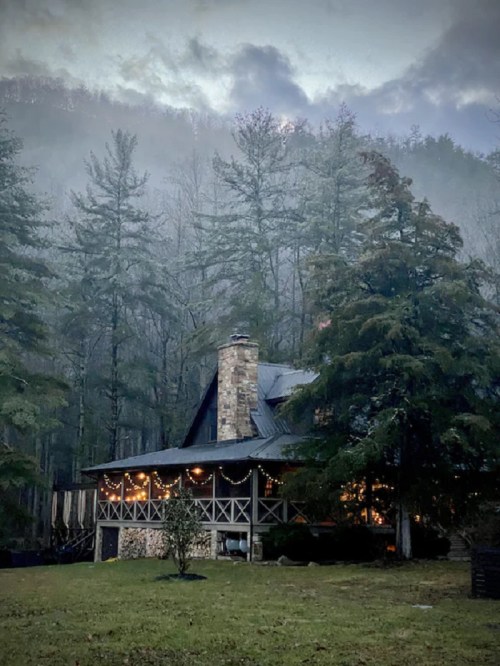 A cozy cabin surrounded by misty trees, adorned with warm lights, set against a backdrop of mountains.