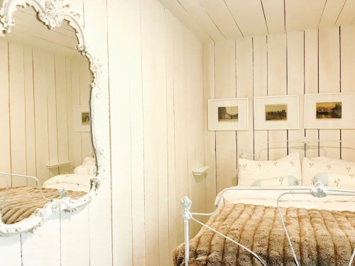 Cozy bedroom with white wooden walls, a vintage mirror, and a plush brown throw on the bed. Framed art hangs nearby.