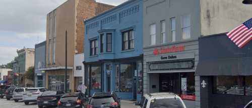 A street view of a small town with a blue building, a State Farm office, and parked cars along the sidewalk.