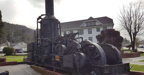 A large, historic steam engine displayed outdoors, with a building and trees in the background under a cloudy sky.