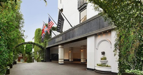 Entrance of a hotel with flags and lush greenery, featuring a fountain and elegant architecture.