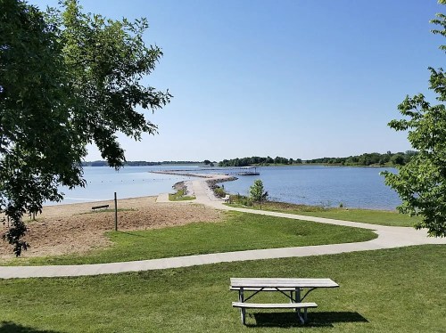 A serene lakeside view with a sandy beach, picnic table, and a pathway leading to a rocky jetty under a clear blue sky.