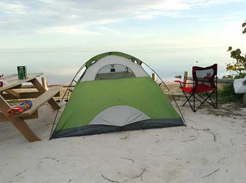 A green tent set up on a sandy beach, with a picnic table and a camping chair nearby, overlooking calm waters.