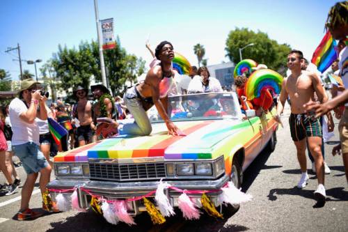 A vibrant parade scene with people celebrating on a rainbow-decorated car, surrounded by colorful flags and joyful participants.
