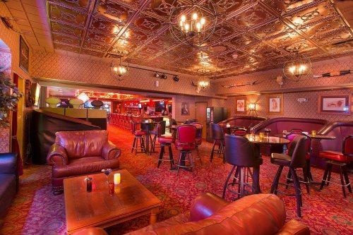 Cozy bar interior with leather seating, ornate ceiling, and dim lighting, featuring tables and a long bar in the background.