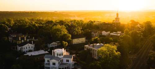 Aerial view of a historic town at sunset, featuring lush trees and classic architecture bathed in golden light.