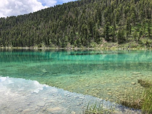 A serene lake with clear turquoise water, surrounded by lush green trees and a mountainous landscape under a cloudy sky.
