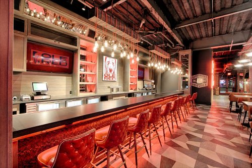 A modern bar interior with a long counter, red lighting, and stylish seating, creating a vibrant atmosphere.