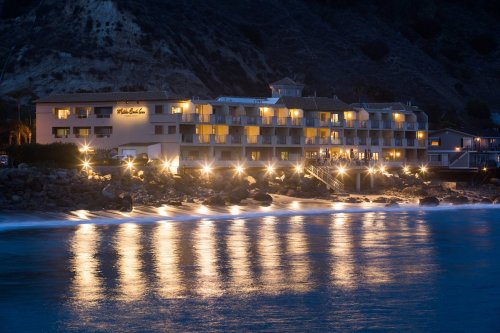 A coastal hotel illuminated at night, reflecting lights on calm water with a rocky shoreline and hills in the background.