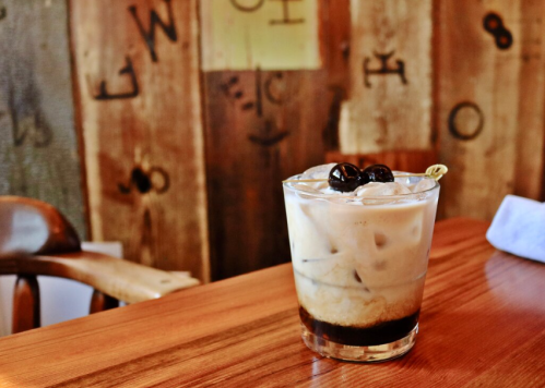 A glass of iced coffee with cream and cherries, placed on a wooden table against a rustic background.