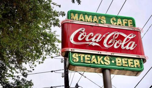 Sign for Mama's Cafe featuring Coca-Cola, with "Steaks" and "Beer" listed below, surrounded by trees and power lines.