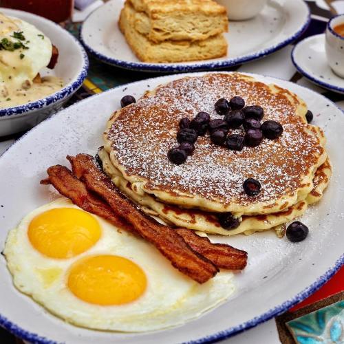 A plate of fluffy pancakes topped with blueberries, served with crispy bacon and two sunny-side-up eggs.