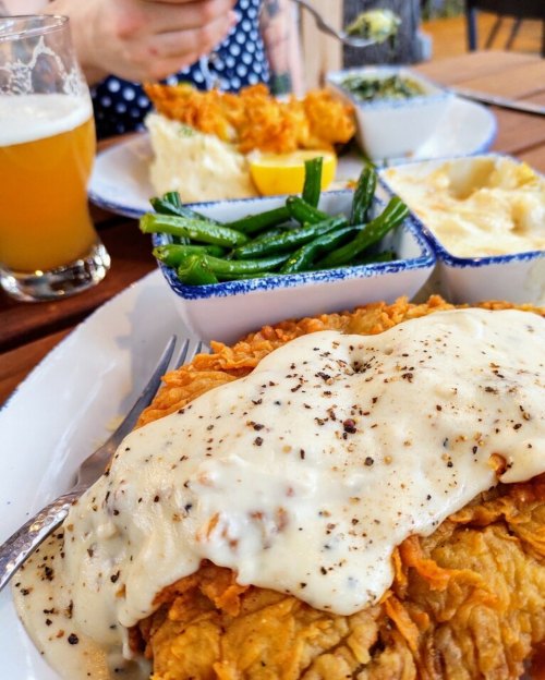 A plate of fried chicken topped with creamy gravy, served with green beans, mashed potatoes, and a glass of beer.