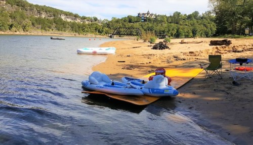 A sandy beach by a lake with inflatable rafts, a chair, and a scenic view of trees and a distant building.