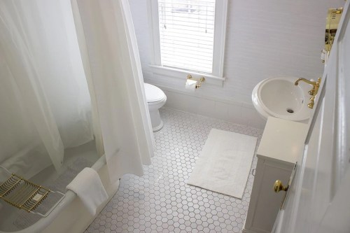 Bright bathroom featuring a shower, toilet, sink, and hexagonal tile flooring with a white rug. Natural light from a window.