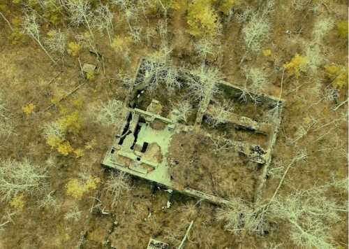 Aerial view of a dilapidated structure surrounded by bare trees and sparse autumn foliage.