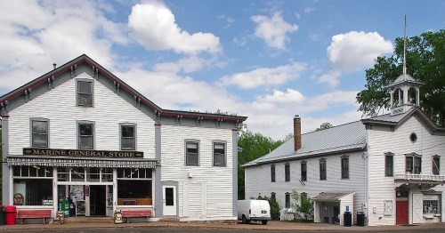 Historic white buildings of the Marine General Store and a nearby structure under a blue sky with fluffy clouds.
