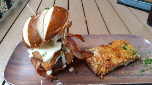 A pretzel bun burger with fried chicken, bacon, and sauce, served alongside a cheesy baked dish on a wooden platter.