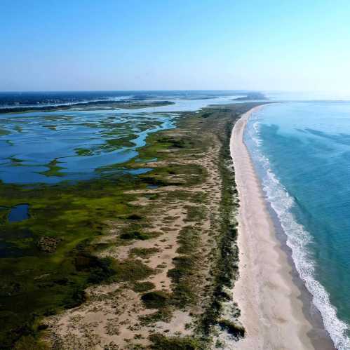 Aerial view of a sandy beach curving along a coastline with lush green wetlands and calm blue waters.