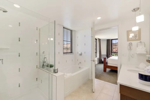 Modern bathroom with a glass shower, bathtub, and a view of a bedroom through an open door. Bright and clean design.
