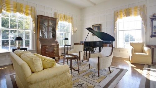 Bright, elegant living room with a grand piano, yellow curtains, and stylish furniture, featuring large windows and wooden floors.