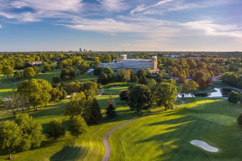 Aerial view of a lush golf course with a hotel, trees, and a city skyline in the background under a clear blue sky.