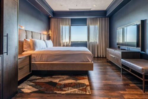 Modern hotel room featuring a king-sized bed, large window, TV, and stylish decor with wooden flooring.