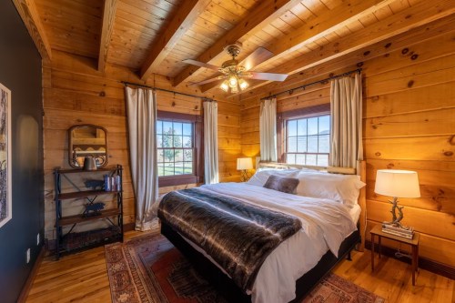 Cozy wooden bedroom with a large bed, warm lighting, and rustic decor, featuring windows and a bookshelf.