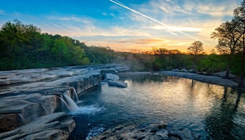 A serene landscape featuring a waterfall flowing into a calm pool, surrounded by lush greenery and a colorful sunset sky.
