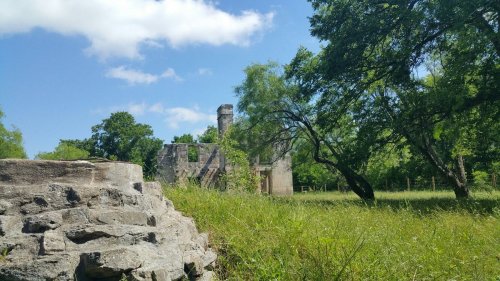 Ruins of a stone building surrounded by lush greenery and trees under a blue sky with scattered clouds.