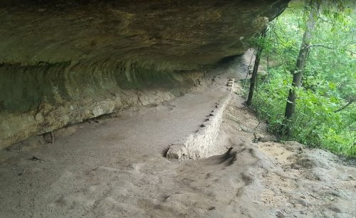 A rocky cave with a smooth ceiling and a dirt path leading through a lush green forest.