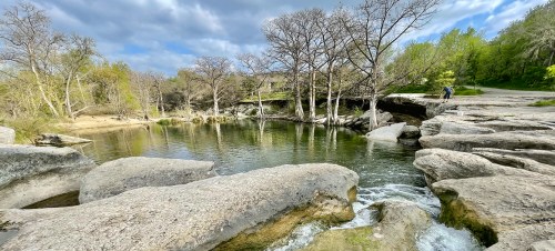 A serene landscape featuring a calm river surrounded by rocky terrain and bare trees under a partly cloudy sky.