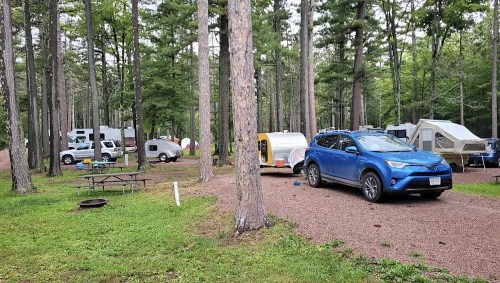 A campsite in a forest with various tents and campers, featuring a blue SUV parked nearby on a gravel path.