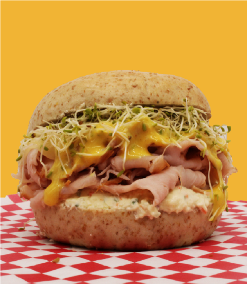 A hearty sandwich with layers of ham, cheese, sprouts, and creamy spread on a whole grain bun, set against a bright yellow background.