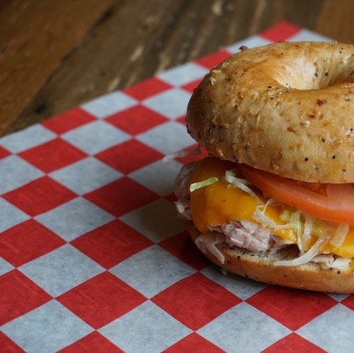 A toasted bagel sandwich filled with turkey, cheddar cheese, lettuce, and tomato on a red and white checkered paper.