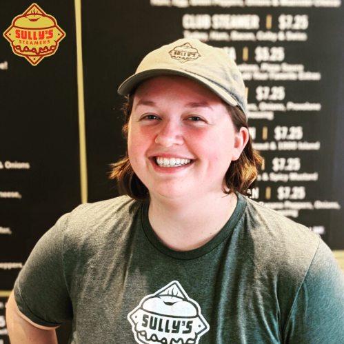 Smiling employee wearing a Sully's Steamers shirt and cap, standing in front of a menu board.