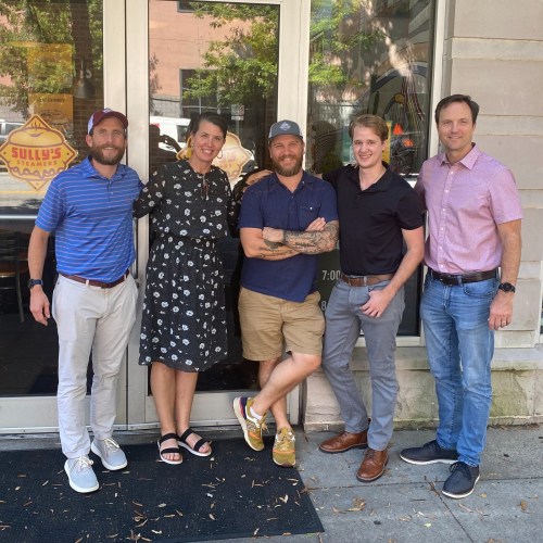 Five people stand outside a restaurant, smiling and posing together in casual attire.