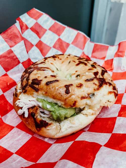 A toasted bagel sandwich with turkey and green spread, served on red and white checkered paper.