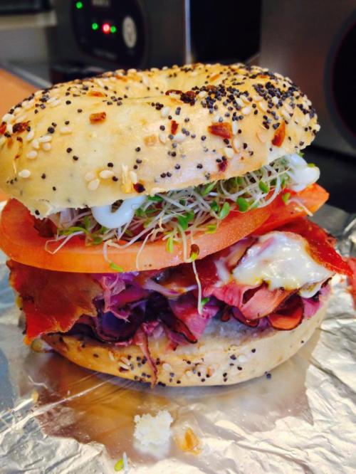 A close-up of a bagel sandwich filled with layers of meats, tomato, sprouts, and mayonnaise, on a foil wrapper.