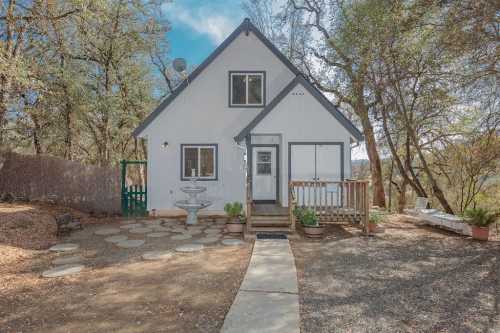 A charming two-story house surrounded by trees, featuring a stone pathway and a small fountain in the front yard.