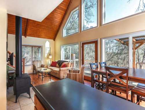 Cozy living space with large windows, wooden beams, and a dining area overlooking a forested view.