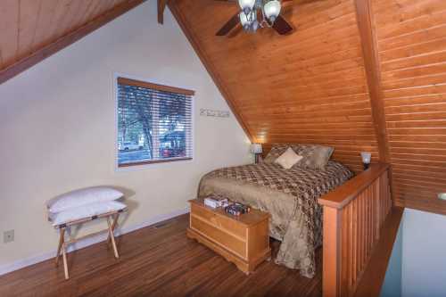 Cozy attic bedroom with wooden walls, a queen bed, and a window overlooking trees. A small table and pillow are nearby.